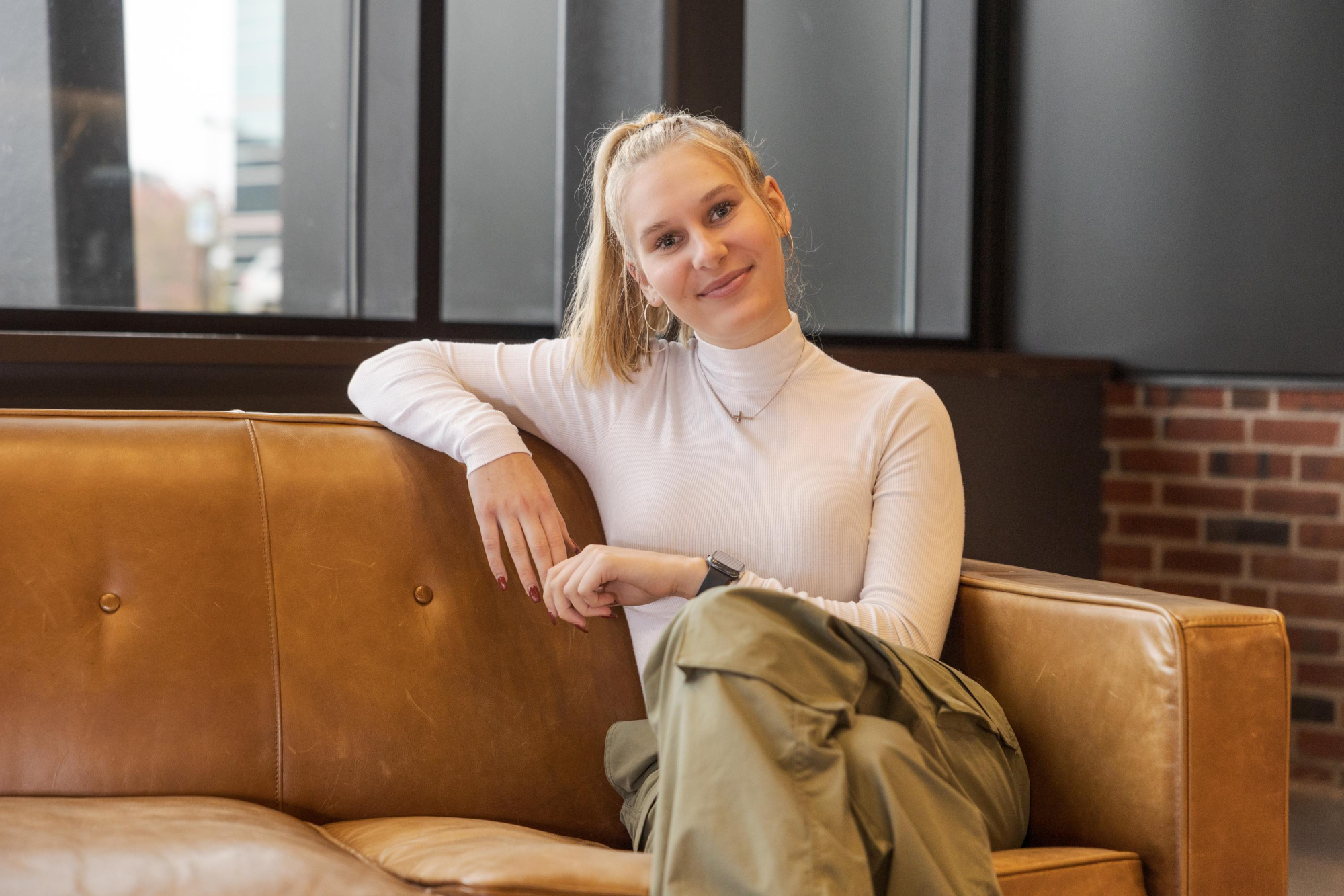 Mackenzie Anderson - A woman with a ponytail, wearing a white turtleneck and green cargo pants, sits on a tan leather couch. She smiles and leans on the armrest. The background includes large windows and brick walls.