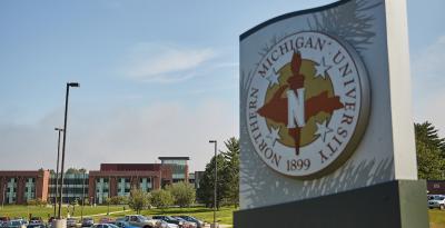 A sign with NMU's seal in the foreground. Jamrich Hall is in the background.